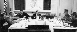 Black and white photo of men sitting around a U-shaped table with an American flag and map of Alaska in the background